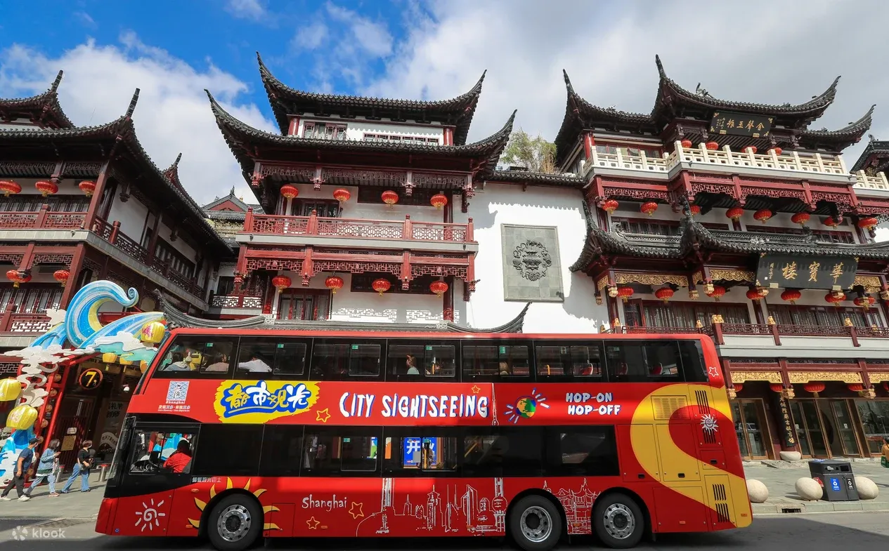 Red double-decker sightseeing bus with "City Sightseeing" branding parked in front of traditional Chinese architecture with curved roofs and red lanterns at Yu Garden
