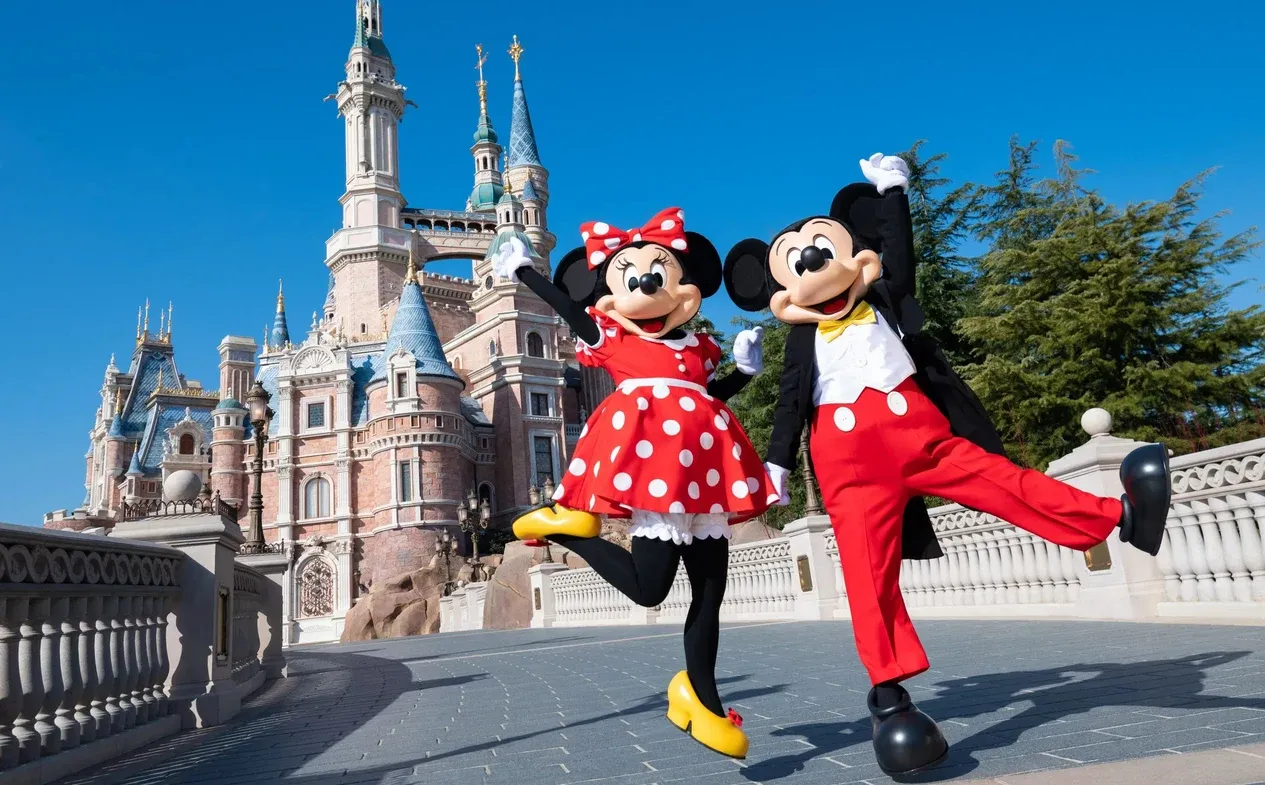 Mickey Mouse in red outfit and Minnie Mouse in polka dot dress standing together in front of pink and blue fairy tale castle with multiple towers and spires