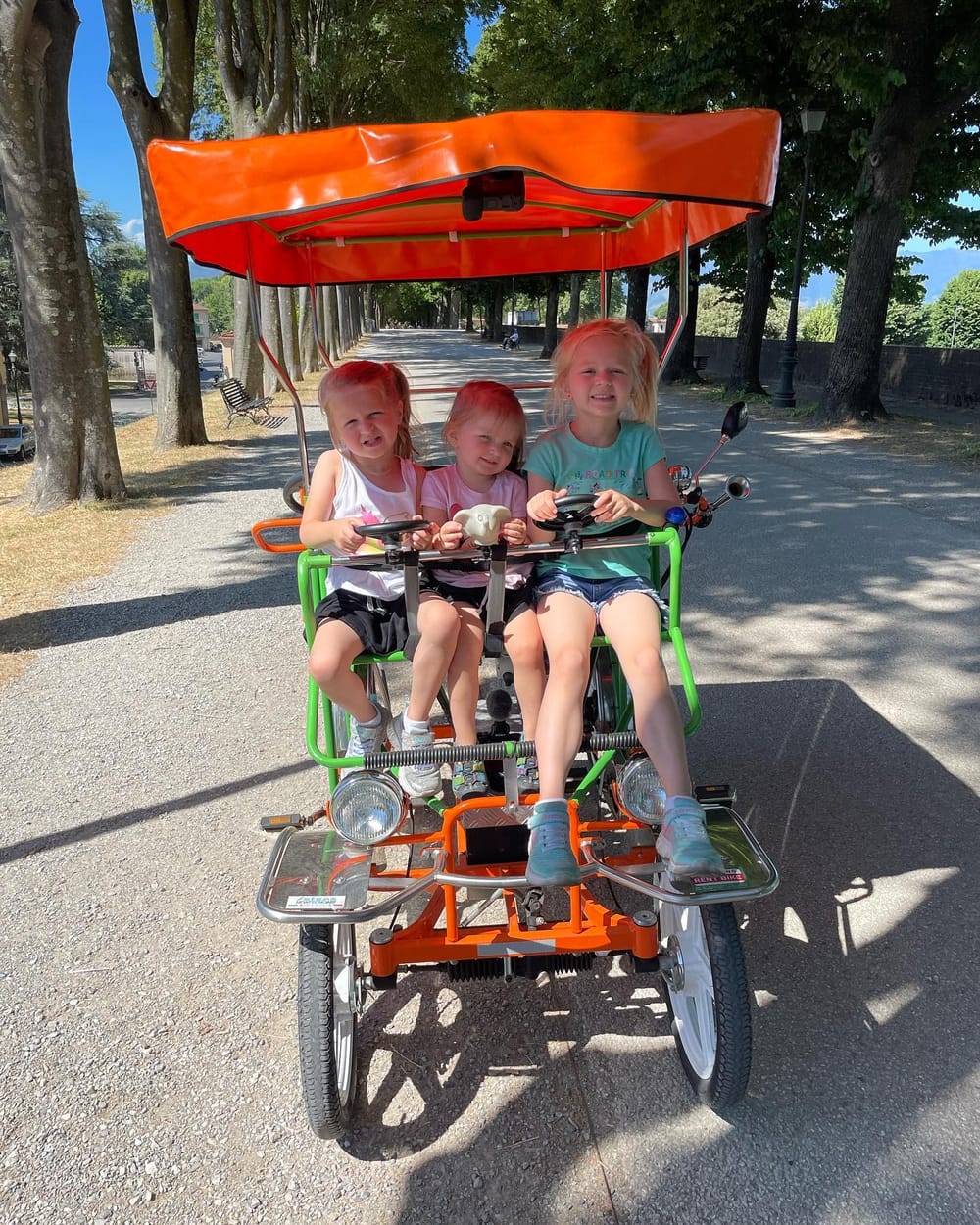 Three girls squeezed side by side across the front seat of an electric pedal car on top of the Lucca city walls